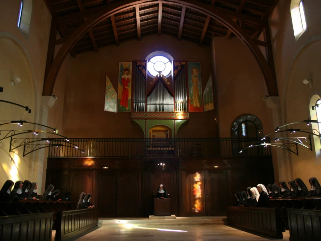 Intérieur d'une chapelle monastique moderne avec des religieuses en prière dans les bancs, un grand orgue à tuyaux et des fresques murales.