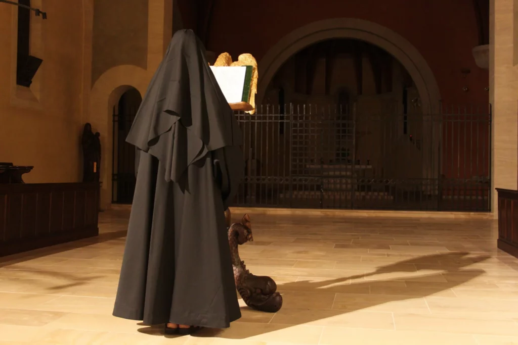 Religieuse en habit noir lisant un livre sur un lectrin aux ailes d'ange, dans une église baignée de lumière.
