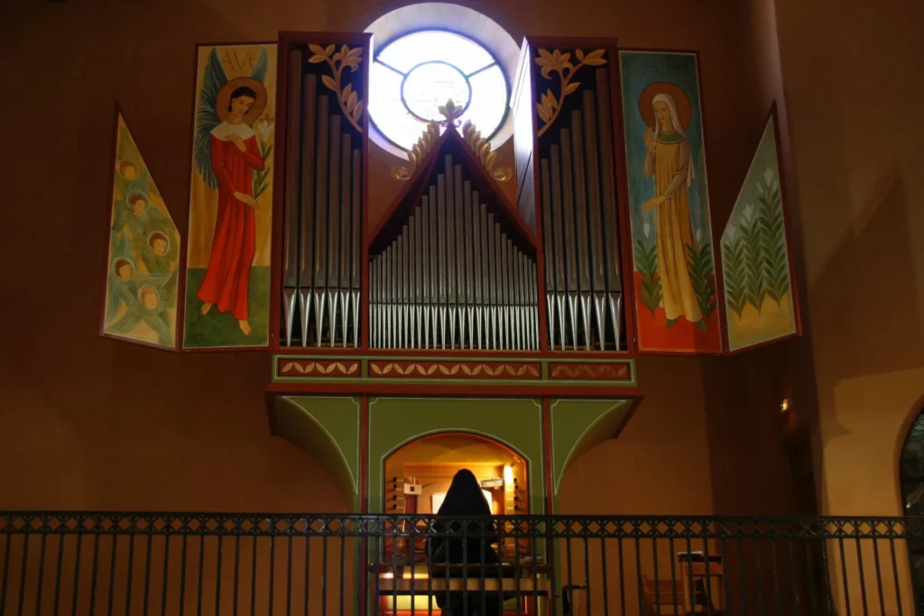 Grand orgue à tuyaux d'église avec des peintures murales de l'Annonciation (Archange Gabriel et Vierge Marie), un vitrail rond, et un organiste en bas.