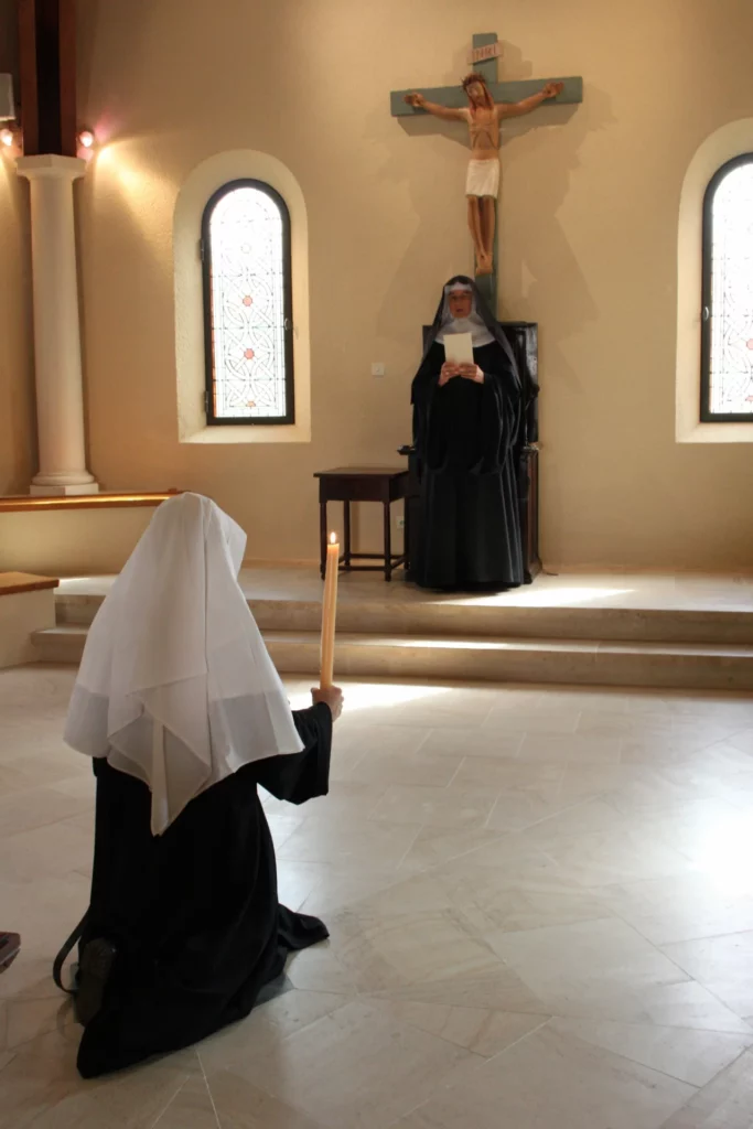 Deux religieuses en habit, l'une agenouillée avec un cierge allumé, l'autre lisant près d'un crucifix dans une église ou chapelle.