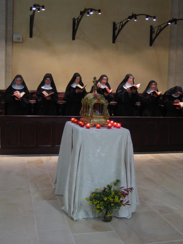 Six moniales en habit religieux lisant un livre dans une chapelle, devant un autel décoré de bougies et d'un reliquaire doré.