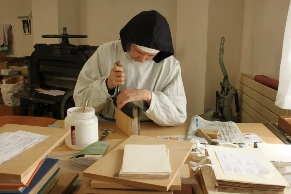 Une religieuse en habit monastique noir et blanc s'appliquant à la reliure artisanale d'un livre ancien dans un atelier traditionnel, entourée de presses et d'outils.