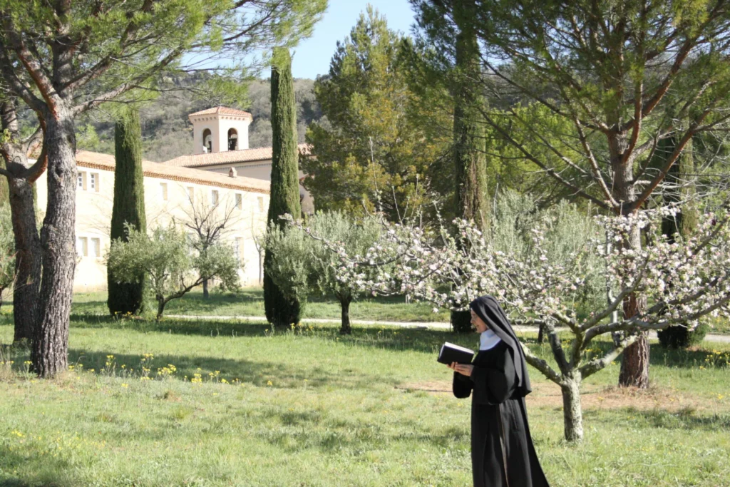 Religieuse en habit noir lisant un livre dans le jardin fleuri d'un monastère en Provence, avec clocher et oliviers.